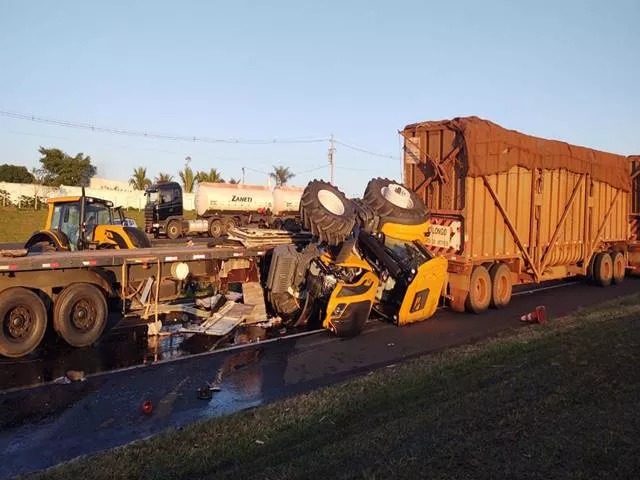 Um caminhão do tipo plataforma conduzindo dois tratores bateu na lateral de um caminhão de cana e os tratores caíram na pista.